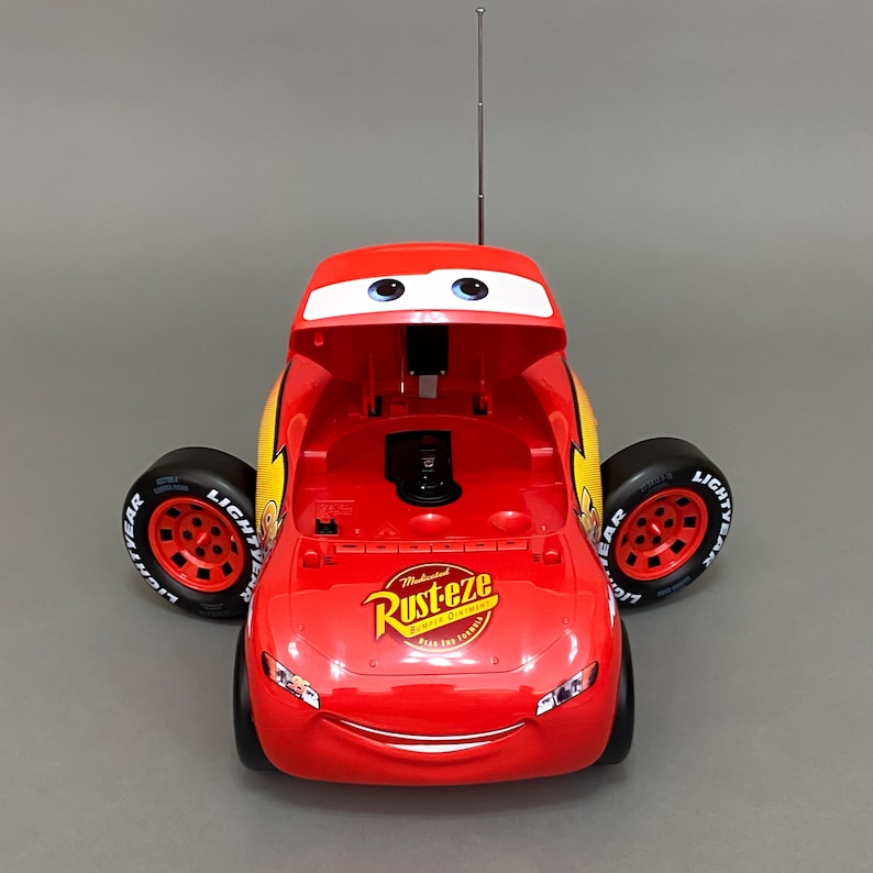 May include: Red toy car shaped like Lightning McQueen from the Disney Pixar Cars movie. The car has a cassette player with the hood open and a Rust-eze sticker on the side.