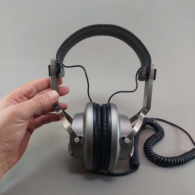 K&ouml;nnte beinhalten: Over-Ear-Kopfh&ouml;rer mit silberfarbenen Ohrmuscheln, schwarzen Polstern, einem dunkelgrauen Kopfb&uuml;gel und sichtbaren Metallverstellarmen. Ein schwarzes Spiralkabel f&uuml;hrt von der Unterseite der Ohrmuscheln weg und erm&ouml;glicht den Kabelanschluss.