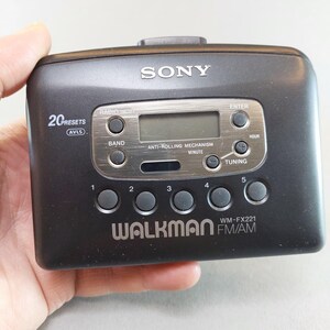 May include: A black Sony Walkman WM-FX221 FM/AM radio cassette player. It features a digital display, control buttons, and the text "WALKMAN" and "SONY". The device is held in a hand.