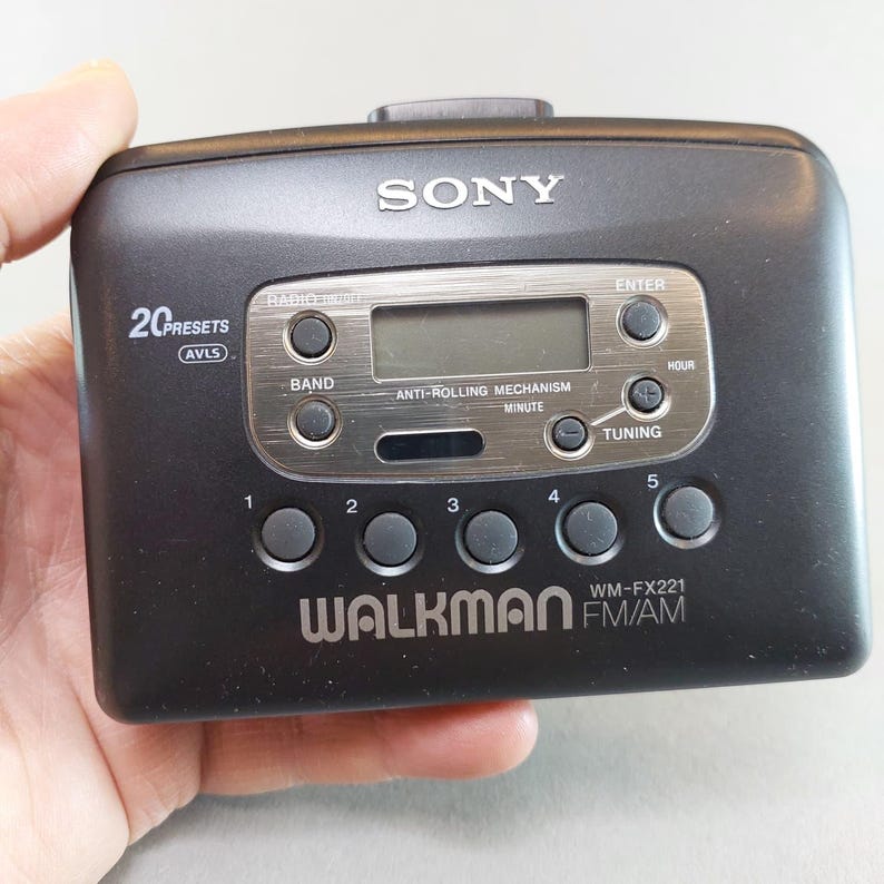 May include: A black Sony Walkman WM-FX221 FM/AM radio cassette player. It features a digital display, control buttons, and the text "WALKMAN" and "SONY". The device is held in a hand.