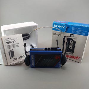 May include: A blue Sony Walkman WM-23 stereo cassette player with black headphones, accompanied by its original blue and white cardboard packaging box, a white styrofoam insert, and a printed instruction manual featuring the model name.