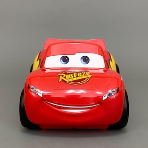 May include: Red toy car with a smiling face and large eyes. The car has a yellow stripe on the side and a logo that says "Rust-eze Bumper Ointment" on the bonnet.