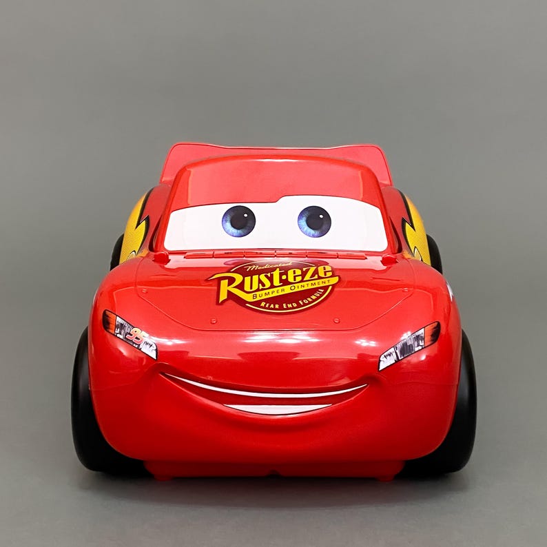 May include: Red toy car with a smiling face and large eyes. The car has a yellow stripe on the side and a logo that says "Rust-eze Bumper Ointment" on the bonnet.