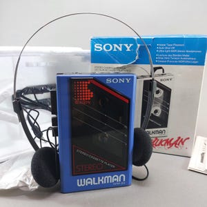 May include: A blue Sony Walkman WM-23 stereo cassette player with a black front panel featuring red graphic accents and the text Sony, Stereo Cassette Player, and Walkman. It includes a thin metal headband with black foam ear pads and its original box.