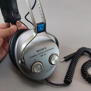 K&ouml;nnte beinhalten: Dynamischer Stereo-Kopfh&ouml;rer Sony DR-S7 in Silber mit schwarzen Ohrpolstern. Die &auml;u&szlig;ere Ohrmuschel verf&uuml;gt &uuml;ber zwei runde Drehregler f&uuml;r Ton und Lautst&auml;rke, eine blaue L-Markierung und ein schwarzes Spiralkabel mit einem Standard-Klinkenstecker.