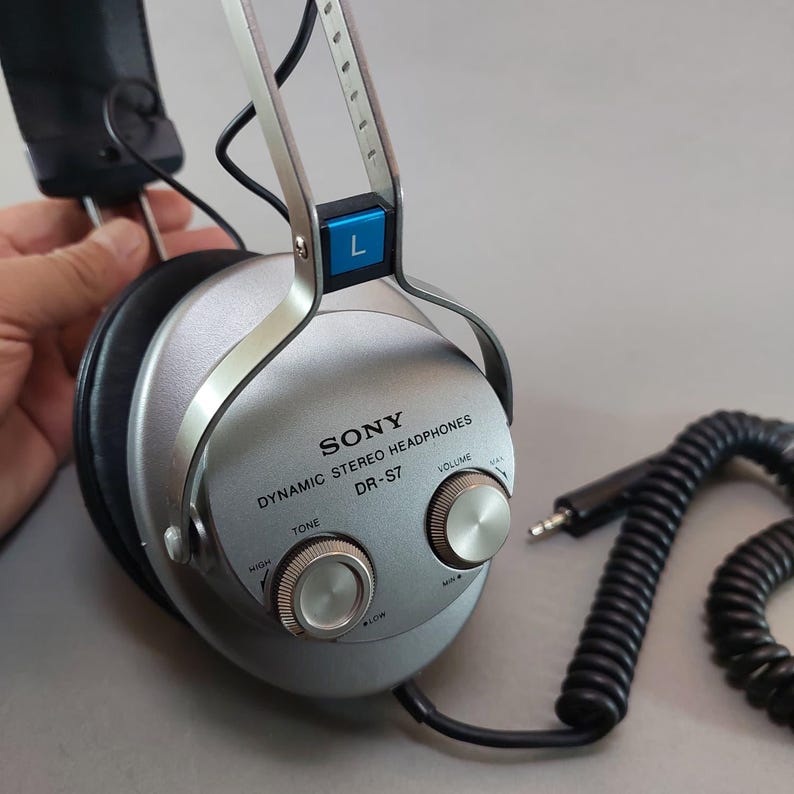 K&ouml;nnte beinhalten: Dynamischer Stereo-Kopfh&ouml;rer Sony DR-S7 in Silber mit schwarzen Ohrpolstern. Die &auml;u&szlig;ere Ohrmuschel verf&uuml;gt &uuml;ber zwei runde Drehregler f&uuml;r Ton und Lautst&auml;rke, eine blaue L-Markierung und ein schwarzes Spiralkabel mit einem Standard-Klinkenstecker.