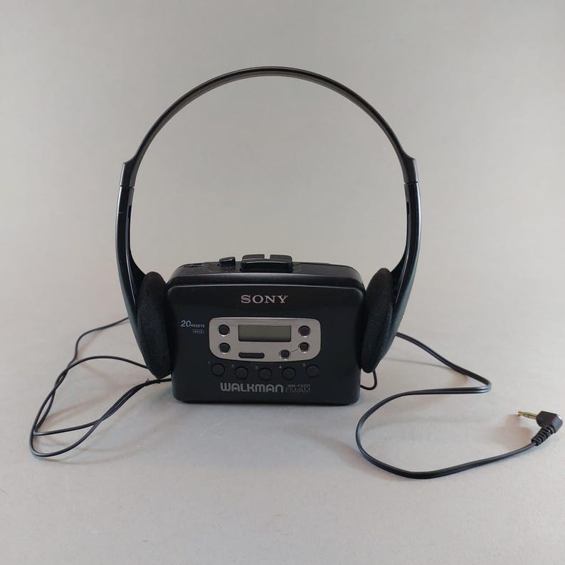May include: A black Sony Walkman cassette player with attached black headphones. The Walkman features a digital display and control buttons. The headphones have black ear pads. The Walkman is connected to the headphones and a cable with a 3.5mm jack.