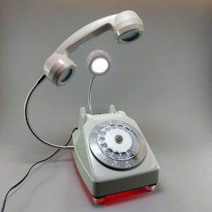 May include: A retro-style telephone lamp in light gray with a rotary dial. The handset and a small lamp are attached with flexible metal arms. The base has red lights. A unique desk lamp with a vintage aesthetic.