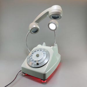 May include: A vintage-style desk lamp in the shape of a rotary phone. The light gray phone has a rotary dial and a handset that arches over the base. The handset and a separate light are illuminated. The base has red lights.