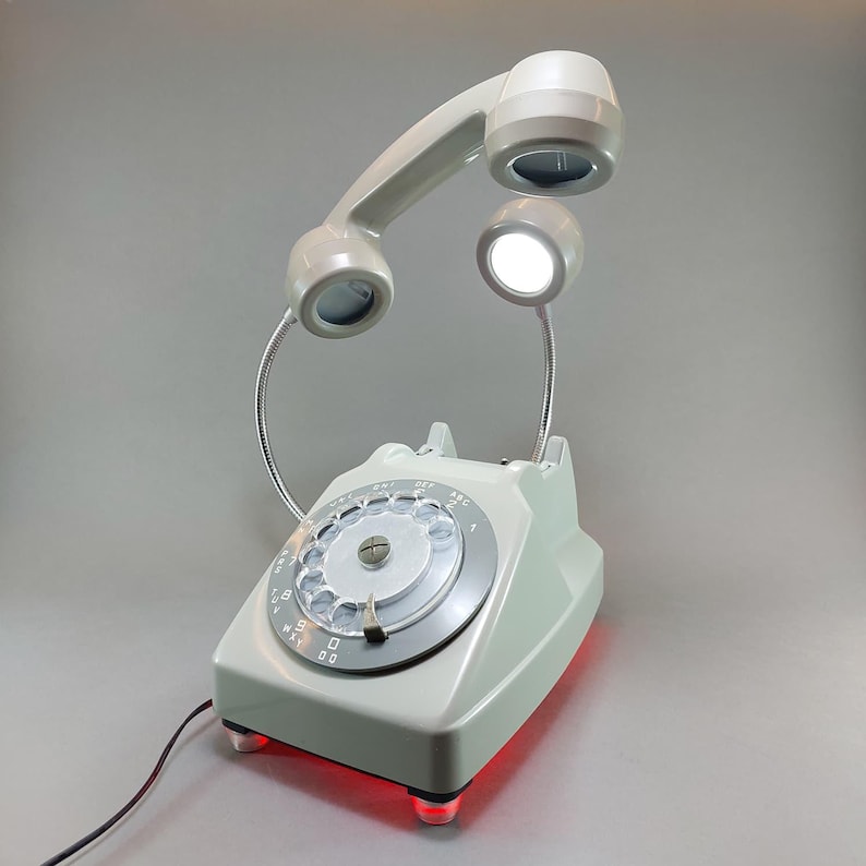 May include: A vintage-style desk lamp in the shape of a rotary phone. The light gray phone has a rotary dial and a handset that arches over the base. The handset and a separate light are illuminated. The base has red lights.