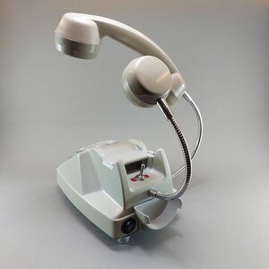 May include: A vintage-style desk lamp in a light gray color, resembling a telephone. The lamp features a handset, a base with a switch, and a flexible metal arm. The design is reminiscent of retro technology.