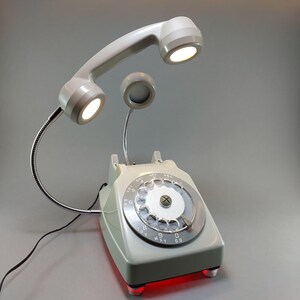 May include: A retro-style desk lamp shaped like a vintage rotary phone. The light gray phone features a dial, a handset with two integrated lights, and a small speaker. The base has a red glow. The lamp is a unique decorative item.