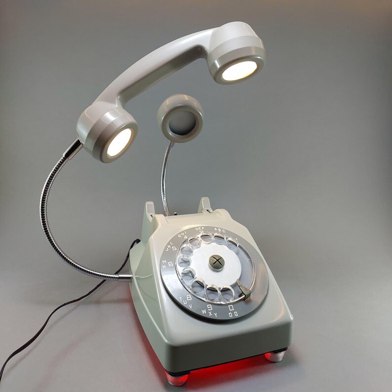 May include: A retro-style desk lamp shaped like a vintage rotary phone. The light gray phone features a dial, a handset with two integrated lights, and a small speaker. The base has a red glow. The lamp is a unique decorative item.