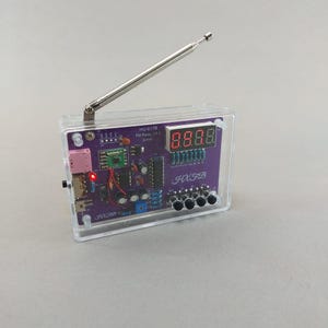 Könnte beinhalten: Ein DIY-UKW-Radio-Bausatz mit einer violetten Leiterplatte in einem transparenten Acrylgehäuse. Das Gerät verfügt über eine silberne Teleskopantenne, eine vierstellige rote LED-Anzeige, vier schwarze Drucktasten und diverse elektronische Bauteile.