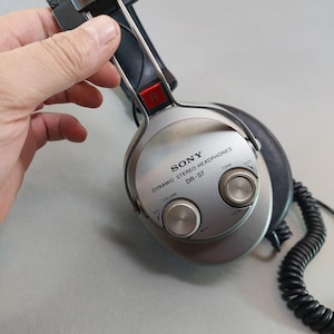 K&ouml;nnte beinhalten: Silberner Sony DR-S7 dynamischer Stereo-Kopfh&ouml;rer mit schwarzem Spiralkabel und schwarzen Ohrpolstern. Die &auml;u&szlig;ere Ohrmuschel zeigt das Sony-Logo, den Modellnamen und zwei runde Drehregler f&uuml;r Lautst&auml;rke und Klang mit einer roten R-Markierung.