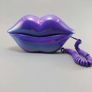 Può includere: Un telefono viola iridescente unico a forma di labbra. Il telefono ha una finitura lucida e un cavo a spirale coordinato. Il design è un pezzo audace, perfetto per aggiungere un tocco di stravaganza a qualsiasi spazio.