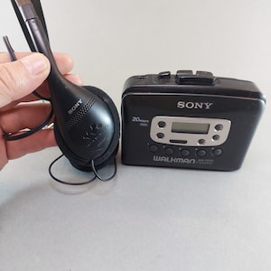May include: Black Sony Walkman cassette player with matching black headphones. The Walkman has a digital display and buttons. The headphones are over-the-ear style. The Walkman is labelled "WALKMAN" and "SONY".