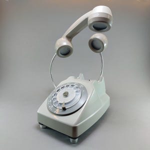 May include: A vintage-style telephone lamp in a soft gray color. The lamp features a rotary dial and a handset that arches overhead, with two circular lights. The base has a rectangular shape with small feet, and the flexible metal arms support the handset.