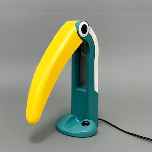May include: A teal and yellow toucan-shaped table lamp with a white accent. The lamp has a round base and a switch on the side.