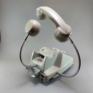 May include: A vintage, light green telephone with a curved handset and a separate earpiece, both connected to the base by flexible, silver-colored tubes. The phone has a rotary dial and a small switch.