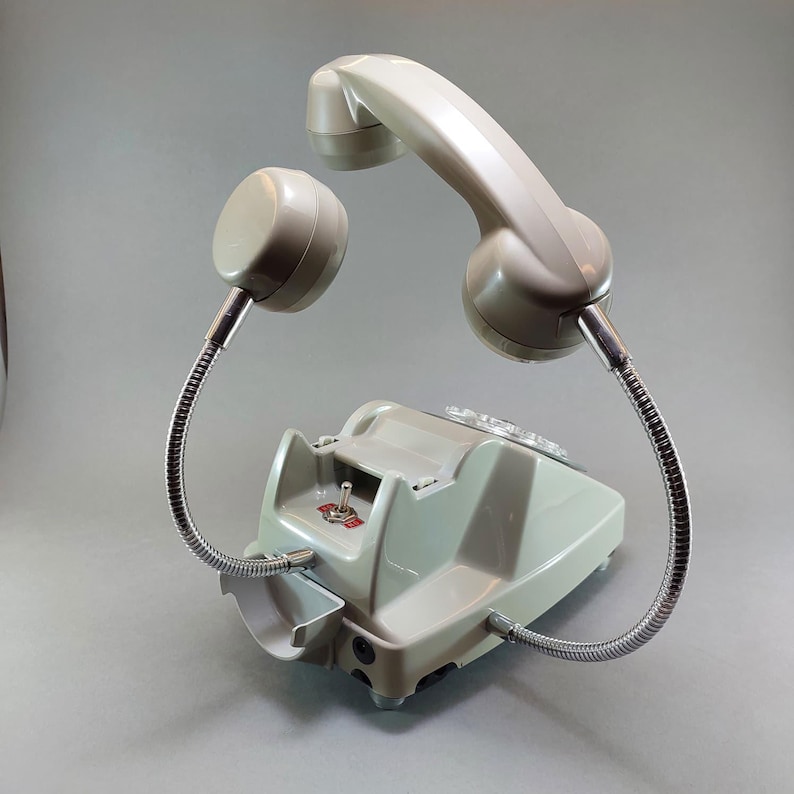 May include: A vintage, light green telephone with a curved handset and a separate earpiece, both connected to the base by flexible, silver-colored tubes. The phone has a rotary dial and a small switch.