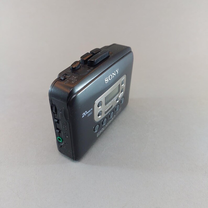 May include: A black Sony Walkman cassette player with silver buttons and a small digital display. The device has a headphone jack and volume controls on the side. The Walkman is a vintage audio player.