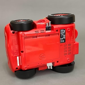 May include: Red toy car with black wheels and a Disney Cars logo on the side. The car is upside down, showing the bottom of the toy. The toy has a battery compartment and a sticker with safety information.
