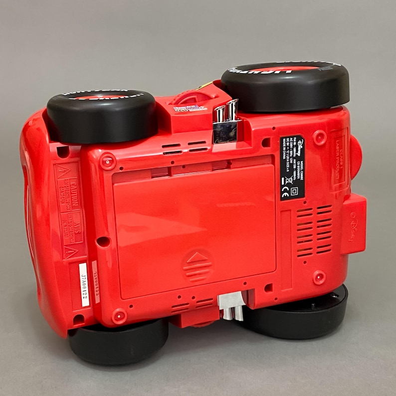 May include: Red toy car with black wheels and a Disney Cars logo on the side. The car is upside down, showing the bottom of the toy. The toy has a battery compartment and a sticker with safety information.