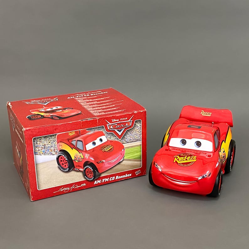 May include: Red and yellow toy car shaped AM/FM CD boombox featuring Lightning McQueen from Disney Pixar Cars. The boombox has a smiling face and the words "RUST-eze" on the side.