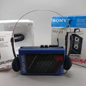 May include: A blue Sony Walkman WM-23 stereo cassette player with black foam headphones and a thin metal headband. The device features black control buttons on top and a clear window. It is accompanied by its original blue and white product box.