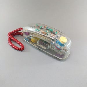 Può includere: Un telefono vintage trasparente che mostra i suoi componenti interni. Ha un cavo a spirale rosso e un pulsante giallo. Il design trasparente rivela i circuiti elettronici, offrendo un'estetica retrò.