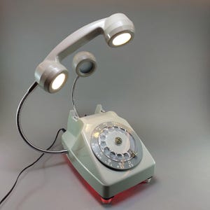 May include: A vintage-style desk lamp in the shape of a rotary phone. The phone is light gray with a rotary dial and a handset that arches overhead. The handset and a separate speaker each have a bright light. The base of the phone glows red.