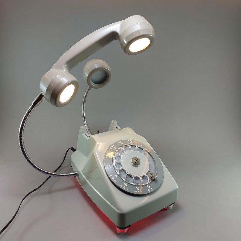 May include: A vintage-style desk lamp in the shape of a rotary phone. The phone is light gray with a rotary dial and a handset that arches overhead. The handset and a separate speaker each have a bright light. The base of the phone glows red.