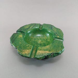 May include: A green ashtray with a speckled pattern and a unique, organic shape. The ashtray has a central bowl and three grooves for resting cigarettes. The color is a vibrant green with yellow accents.