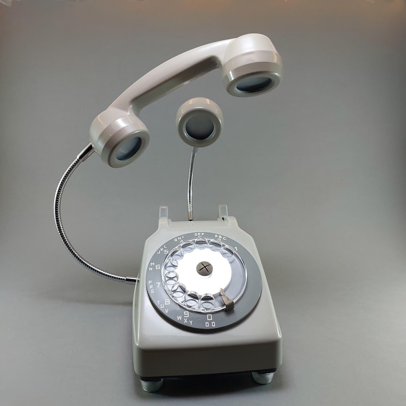 May include: A gray vintage rotary phone converted into a desk lamp. The phone's handset and earpiece are positioned above the base, with a flexible metal arm. The dial features numbers and letters, and the lamp emits a soft glow.