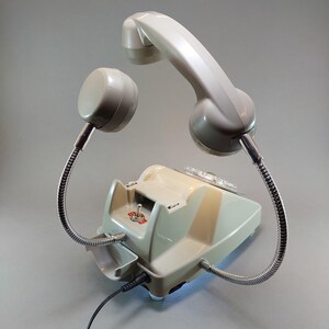 May include: A vintage-style telephone in a light gray color scheme. The handset is off the hook, with a flexible metal tube connecting it to the base. The base has a rotary dial and a switch.