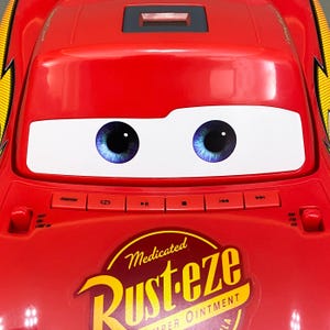 May include: Red toy car shaped like Lightning McQueen from the Disney Pixar Cars movie. The car has a cassette player with buttons for play, rewind, fast forward, and stop. The car has a sticker that says "Medicated Rust-eze Super Ointment".
