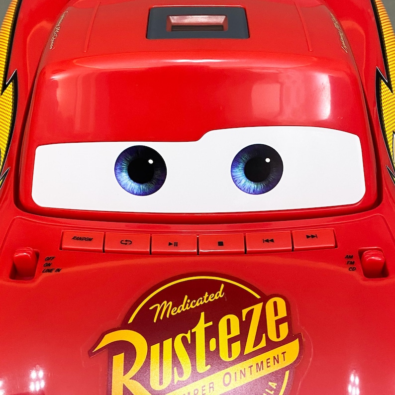 May include: Red toy car shaped like Lightning McQueen from the Disney Pixar Cars movie. The car has a cassette player with buttons for play, rewind, fast forward, and stop. The car has a sticker that says "Medicated Rust-eze Super Ointment".
