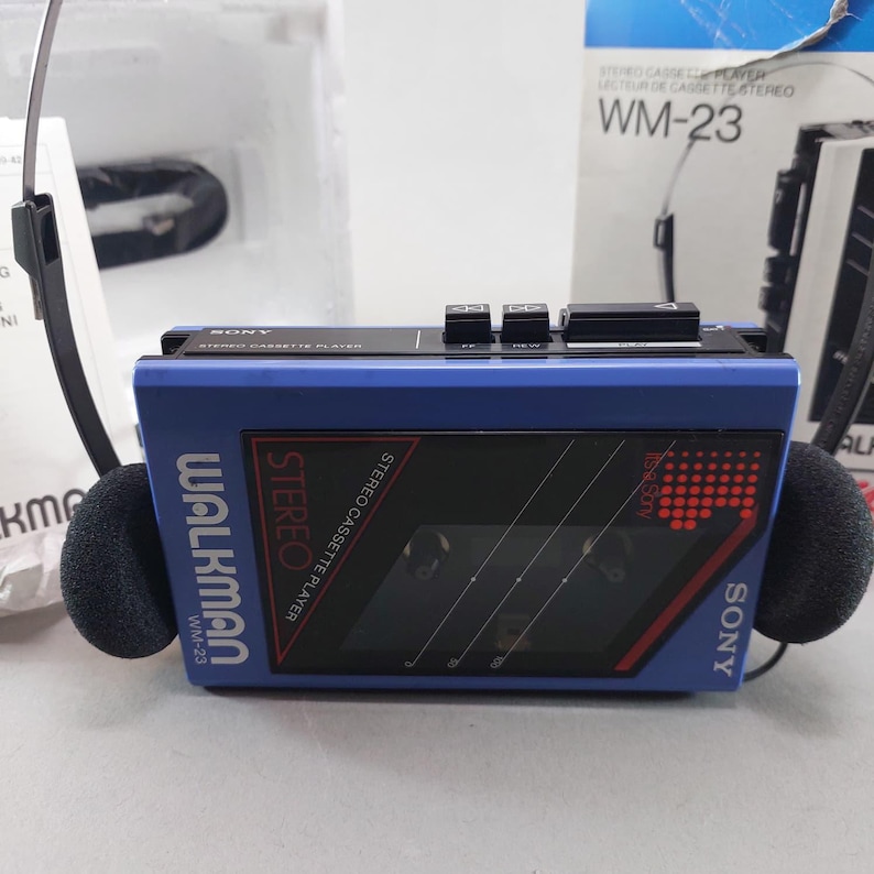 May include: A blue Sony Walkman WM-23 stereo cassette player with black control buttons on top, a transparent cassette window, and black foam-covered headphones attached. The device features white and red text including Walkman and Stereo.