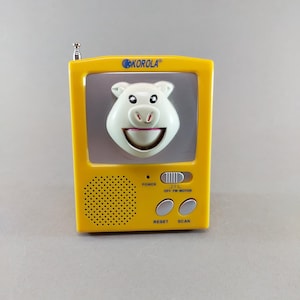 Könnte beinhalten: Ein gelbes tragbares Radio mit einem schweineförmigen Gesicht auf dem Bildschirm. Das Radio hat eine silberne Antenne, den Markennamen "KOROLA" und Tasten für Ein/Aus, Reset und Scan. Das Schwein hat einen lächelnden Mund und kleine Augen.