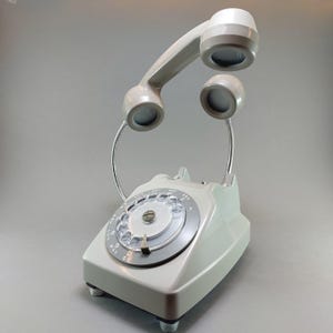 May include: A vintage-style telephone lamp in a light gray color. The lamp features a rotary dial and a handset that arches overhead, with two smaller lights on flexible metal stems. The base has a rectangular shape with four small feet.
