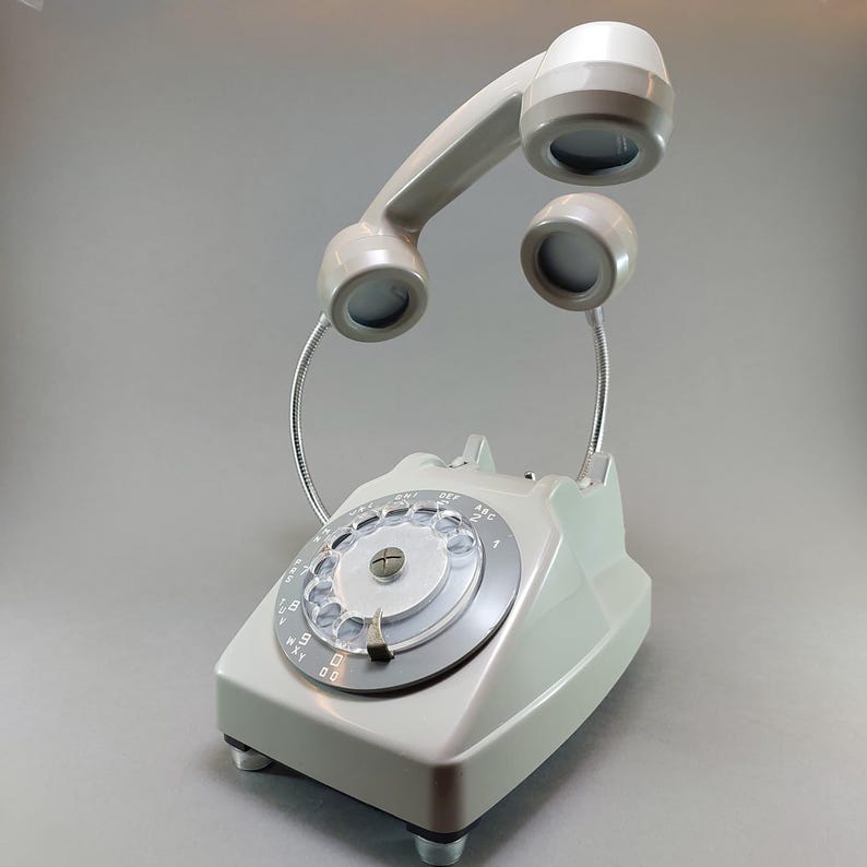 May include: A vintage-style telephone lamp in a light gray color. The lamp features a rotary dial and a handset that arches overhead, with two smaller lights on flexible metal stems. The base has a rectangular shape with four small feet.