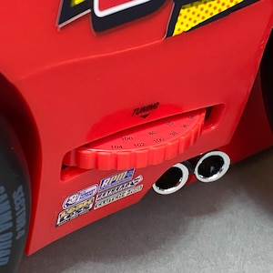 May include: Red toy car with a black and yellow stripe design. The car has a tuning knob with numbers from 70 to 104. The car has two chrome exhaust pipes.