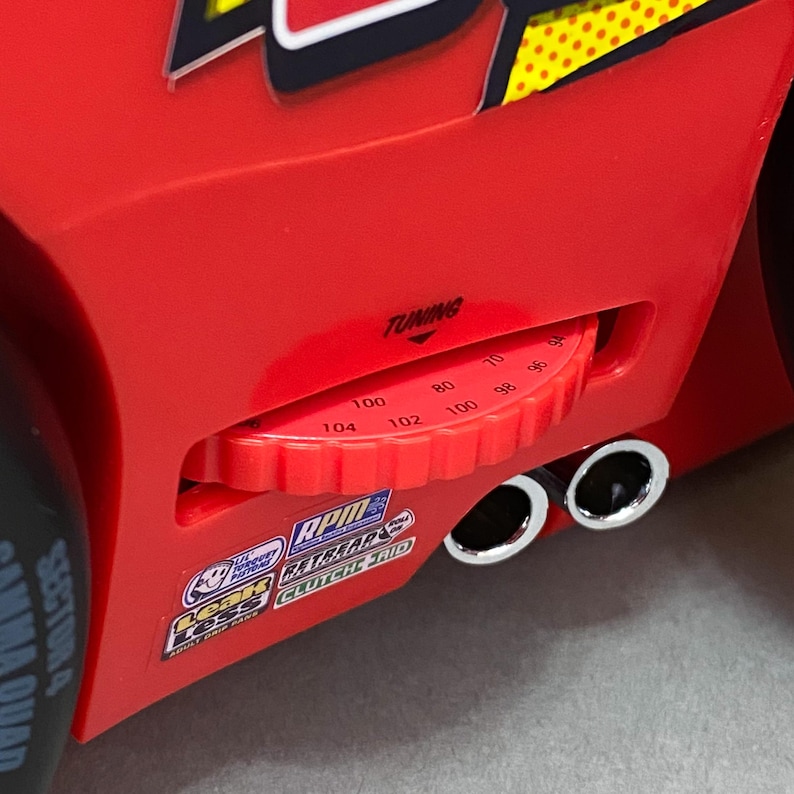 May include: Red toy car with a black and yellow stripe design. The car has a tuning knob with numbers from 70 to 104. The car has two chrome exhaust pipes.