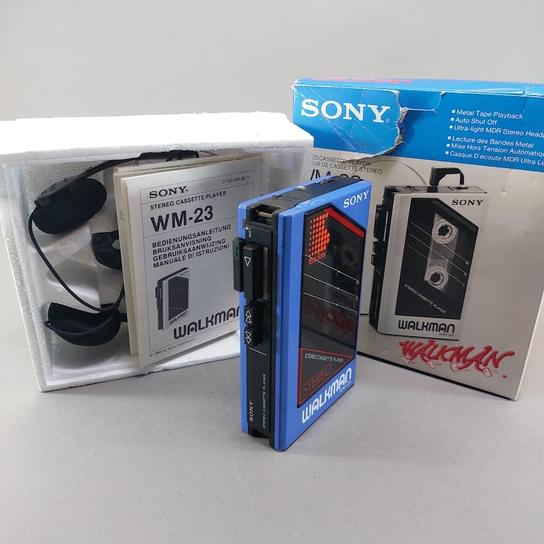 May include: A blue Sony Walkman WM-23 stereo cassette player with black control buttons, accompanied by black headphones, a user manual, and the original blue and white cardboard packaging box featuring product specifications and branding.