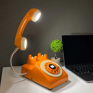 May include: Orange rotary phone shaped desk lamp with a flexible arm and two white light bulbs. The phone has a white dial and a white receiver.