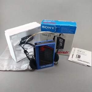 May include: A blue Sony Walkman WM-23 cassette player with black headphones, original blue and white packaging box, white styrofoam insert, and a printed instruction manual. The device features the Sony logo and Walkman branding on the front.