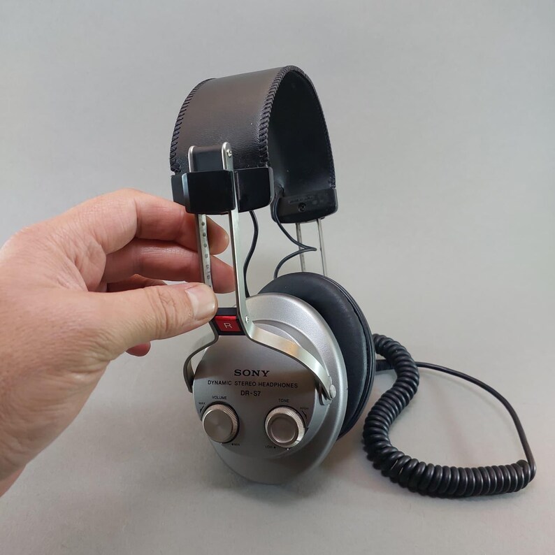 K&ouml;nnte beinhalten: Vintage Sony DR-S7 dynamischer Stereo-Kopfh&ouml;rer mit dunkelbraunem gepolstertem Kopfb&uuml;gel, silbernen Ohrmuscheln mit integrierten Lautst&auml;rke- und Klangreglern sowie einem schwarzen Spiralkabel. Die rechte Seite ist mit einem roten R markiert.