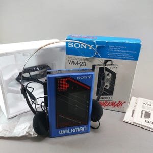 May include: A blue Sony Walkman WM-23 stereo cassette player with black accents, accompanied by a pair of black foam-padded headphones, the original blue and white product box, white styrofoam packaging, and a printed instruction manual.