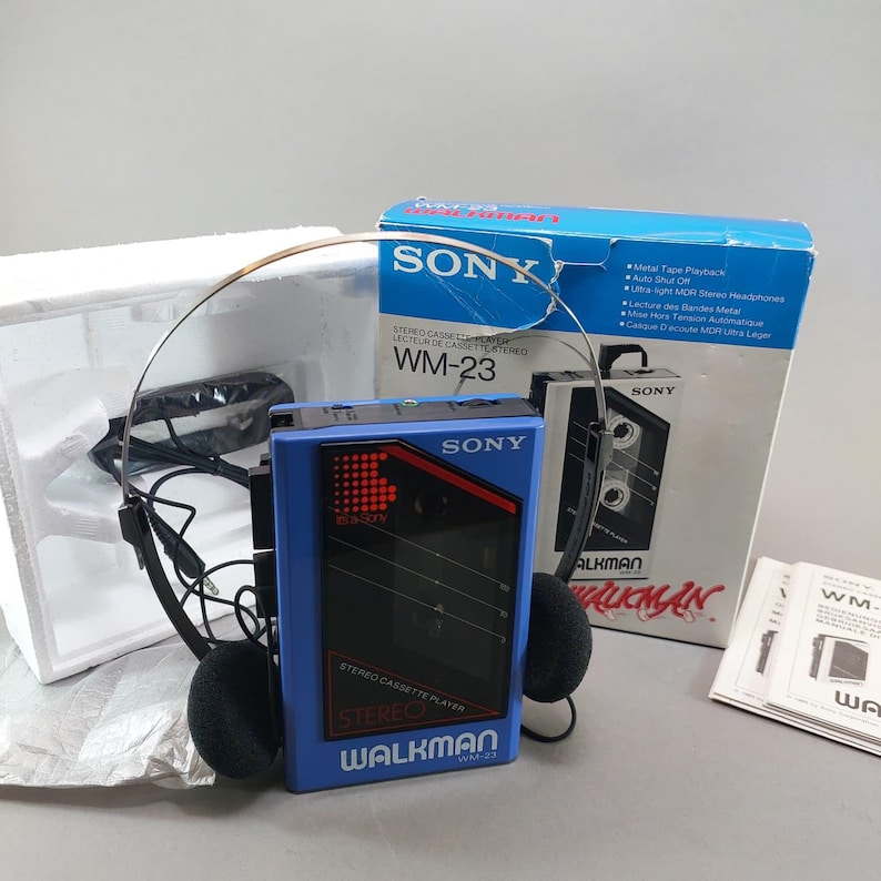 May include: A blue Sony Walkman WM-23 stereo cassette player with black accents, accompanied by a pair of black foam-padded headphones, the original blue and white product box, white styrofoam packaging, and a printed instruction manual.